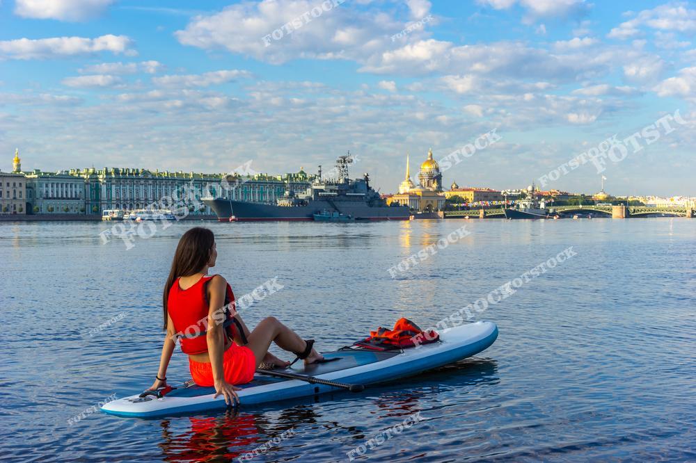 На переднем плане изображена девушка на сапборде, отдыхающая на поверхности Невы. На заднем плане — Зимний дворец, входящий в ансамбль Эрмитажа, купол Исаакиевского собора и военное судно у набережной. Также видны шпиль Адмиралтейства и Троицкий мост. Ясное небо и отражения в воде создают атмосферу спокойствия и гармонии. Это типичный летний пейзаж Санкт-Петербурга, сочетающий активный отдых и величие архитектуры.
